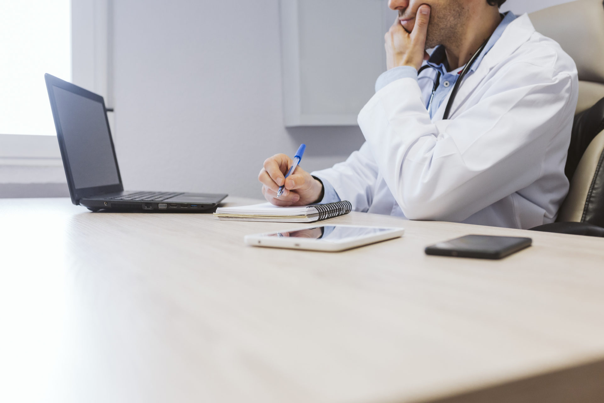 Midsection Of Doctor Writing In Note Pad By Laptop On Desk